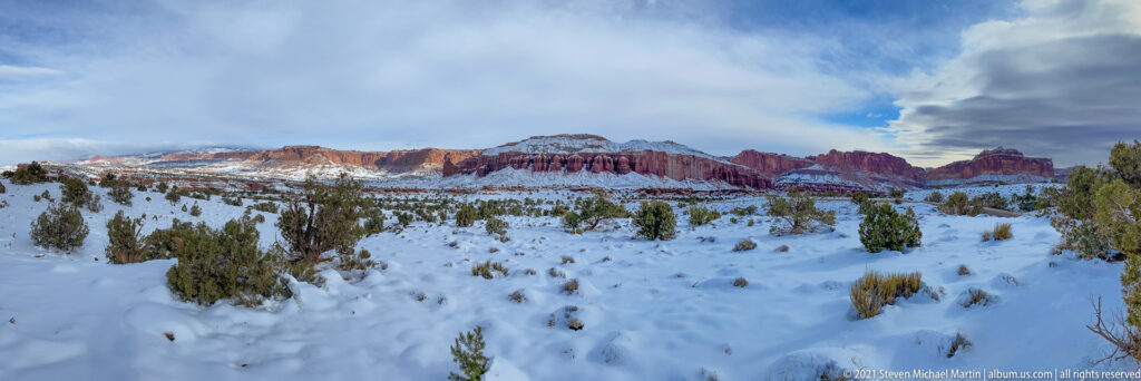 Snowy ridges and layered cliffs extend beneath clouds, with scattered junipers across the foreground.