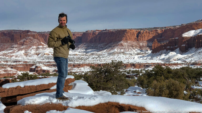 A man holding a camera stands on a snowy rock ledge above a wide red-rock canyon.