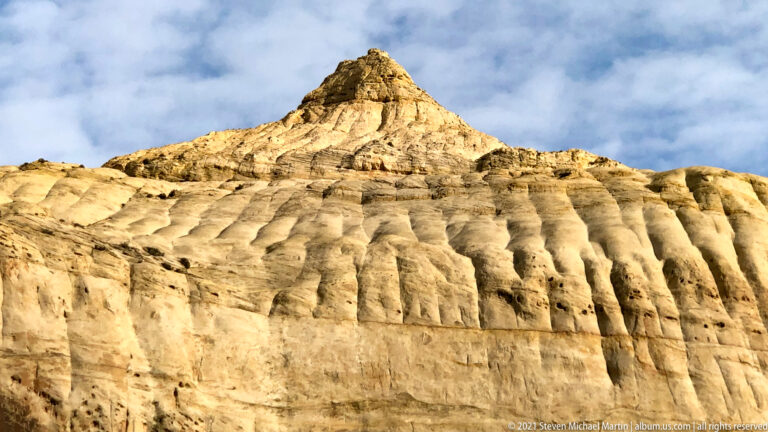 A pointed pale sandstone summit rises over a ribbed cliff beneath a cloudy blue sky.