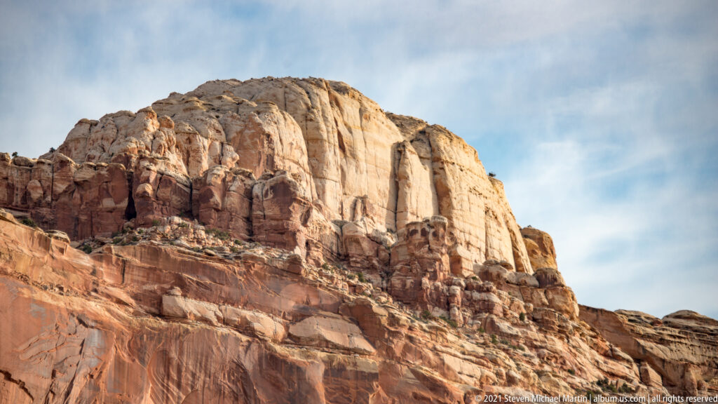 A tall pale cliff with vertical seams rises above red layered ledges beneath a cloudy sky.