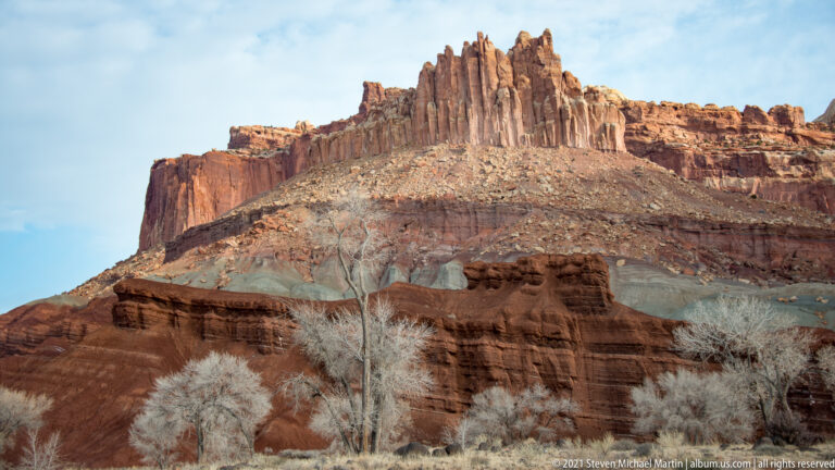 Narrow sandstone fins crown a red cliff above a pale slope and bare trees.