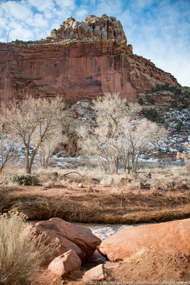 Bare shrubs and snow patches fill a wash beneath a long red cliff capped with pale rock.