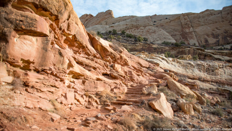 Stone steps ascend a rocky passage between sandstone walls and scattered shrubs.
