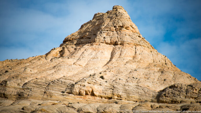 A pointed pale sandstone summit rises above folded horizontal layers beneath blue sky.