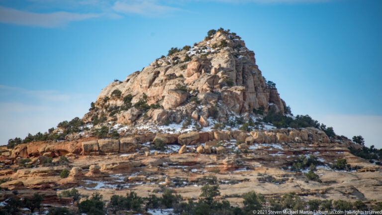 A rounded sandstone dome with junipers and snow patches rises above red layered ledges.