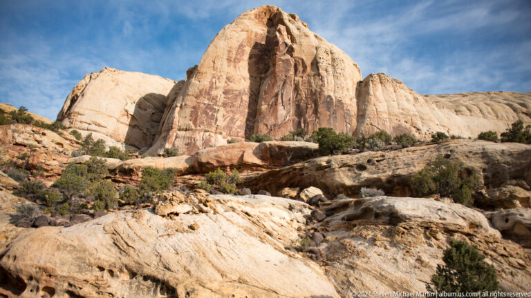 Tall pale sandstone monoliths with dark streaks rise above broken ledges and shrubs.