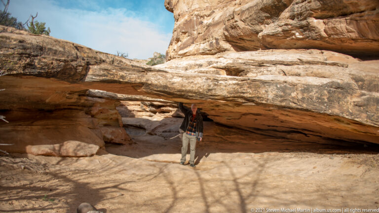 A person stands inside a broad shaded sandstone shelter beneath a low projecting ceiling.