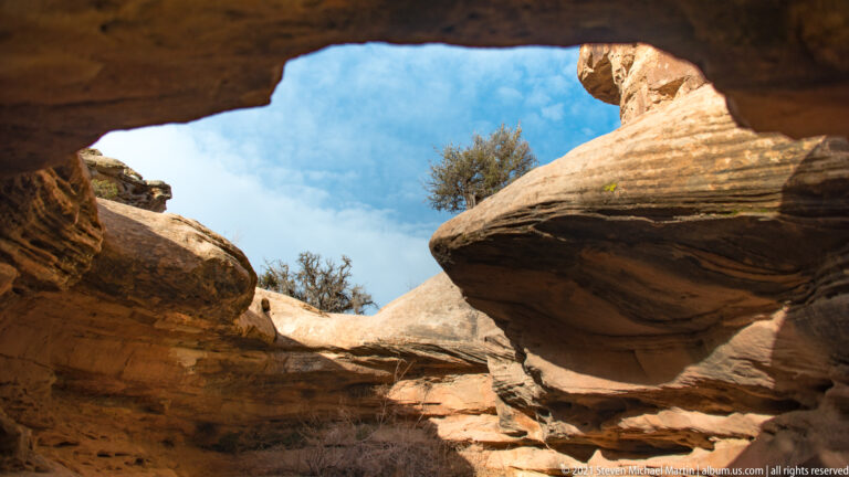 A gap between shadowed sandstone ledges frames blue sky, shrubs, and sunlit rock.