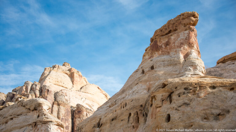 A slender sandstone pinnacle with a projecting cap rises against blue sky above pale rocks.