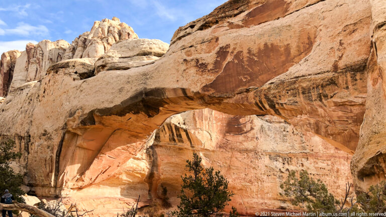 A broad sandstone bridge spans a shaded alcove, with a hiker at the lower left for scale.