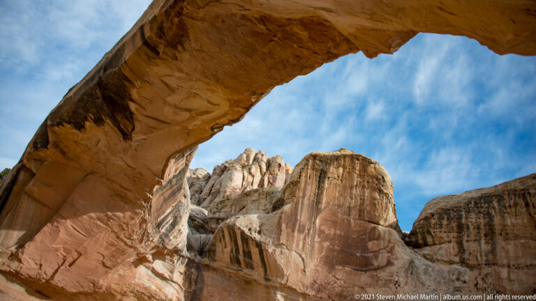 The shaded underside of Hickman Bridge arches across the top of the view, framing pale cliffs and blue sky.