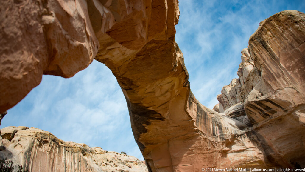 The bridge spans overhead between steep sandstone walls, with a pale cliff and blue sky visible through the opening.