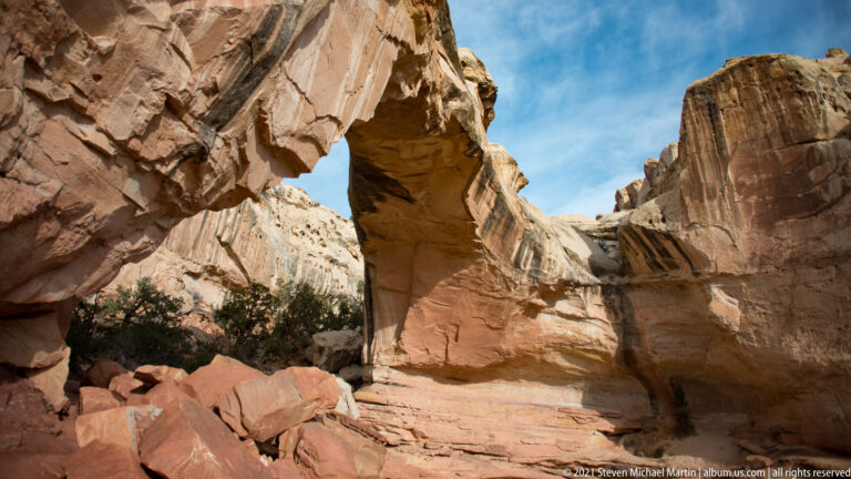 A red sandstone arch frames pale cliffs beyond, above broken rocks and sparse shrubs.