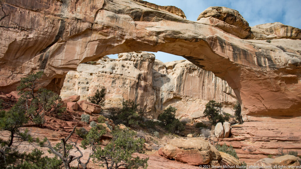 A red sandstone bridge spans above shrubs and broken rock, framing a pale cliff through its opening.