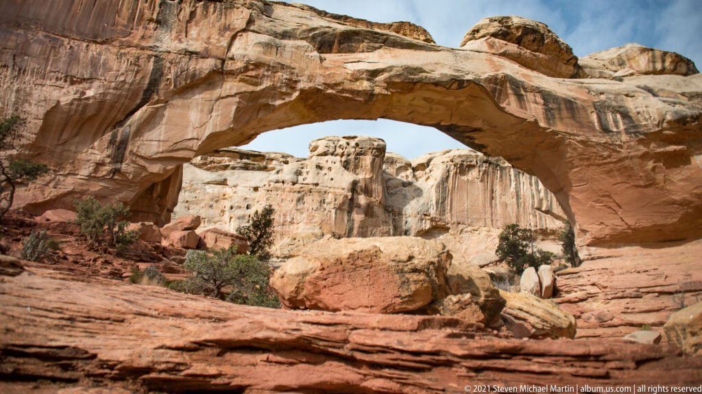 A large sandstone boulder sits below Hickman Bridge, with pale cliffs and sparse shrubs beyond.