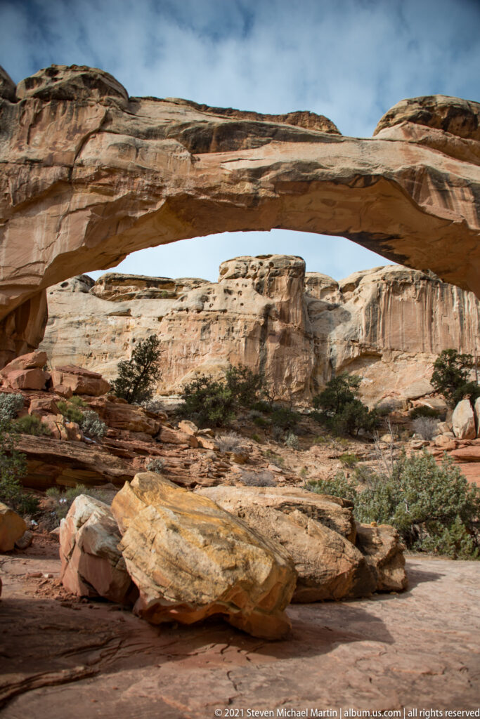 Hickman Bridge spans a broad canyon above layered sandstone slabs and shrubs under a partly cloudy sky.