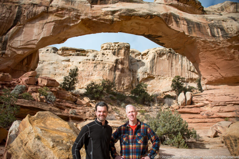 Two men pose beneath Hickman Bridge, with pale cliffs framed by the sandstone arch behind them.