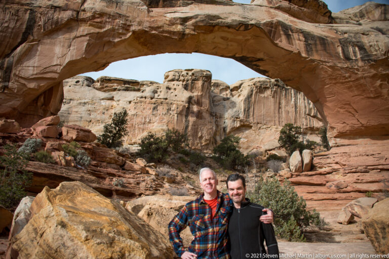 Two men stand beside a large boulder beneath Hickman Bridge, with pale canyon cliffs behind them.