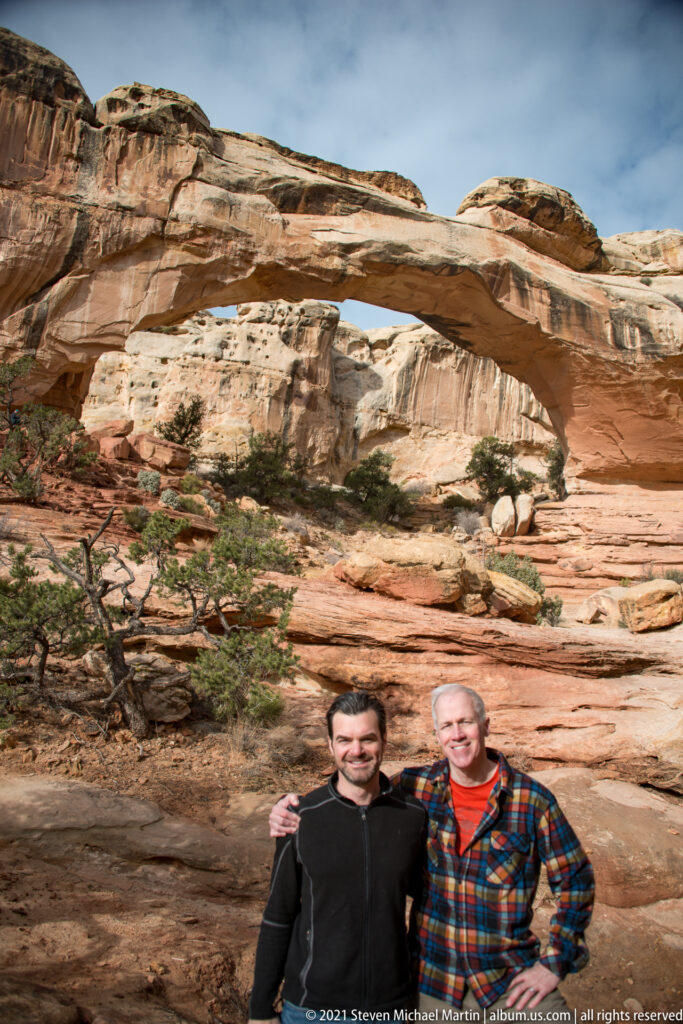 Two men smile beneath Hickman Bridge, with the broad sandstone span and cliffs above them.