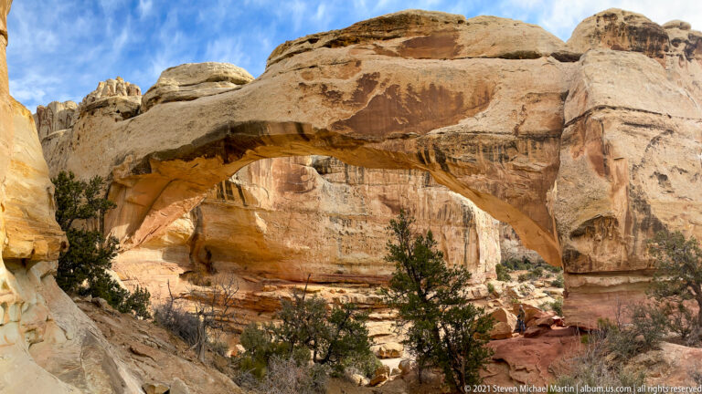 A small visitor stands below Hickman Bridge, which spans a pale sandstone bowl dotted with shrubs.