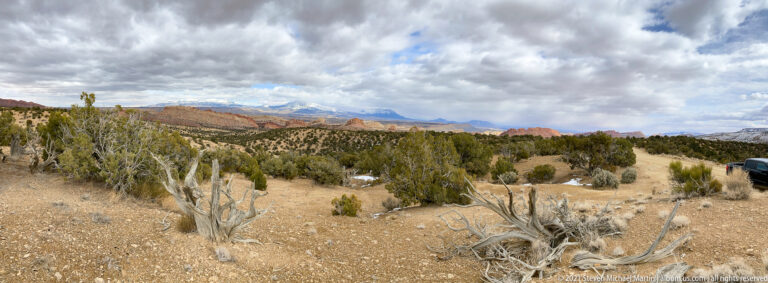 Juniper-dotted plateau with distant layered cliffs, snow patches and part of a vehicle at right.