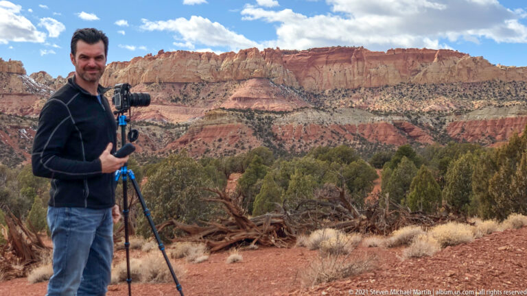 A man stands beside a camera on a tripod with red and cream canyon cliffs behind him.