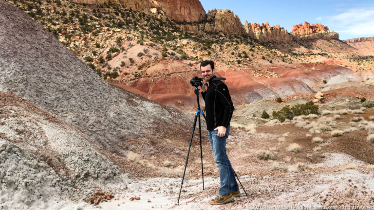 A man stands beside a tripod on pale rock, with layered red sandstone cliffs behind him.