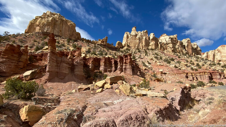 Pale sandstone towers line a ridge above darker horizontal red ledges and scattered boulders.
