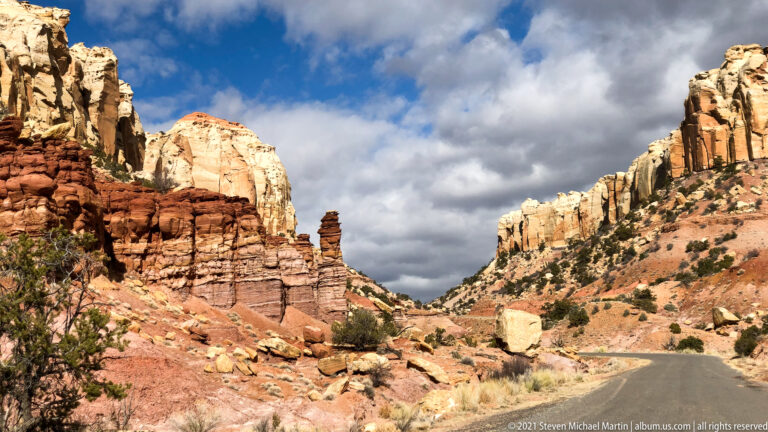 A paved road curves between dark red ledges and pale sandstone towers under a partly cloudy sky.