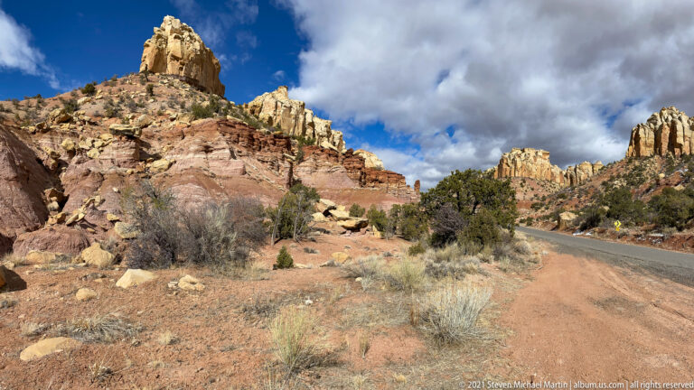 A paved road curves beside layered red and cream cliffs, with grasses and a small roadside sign.