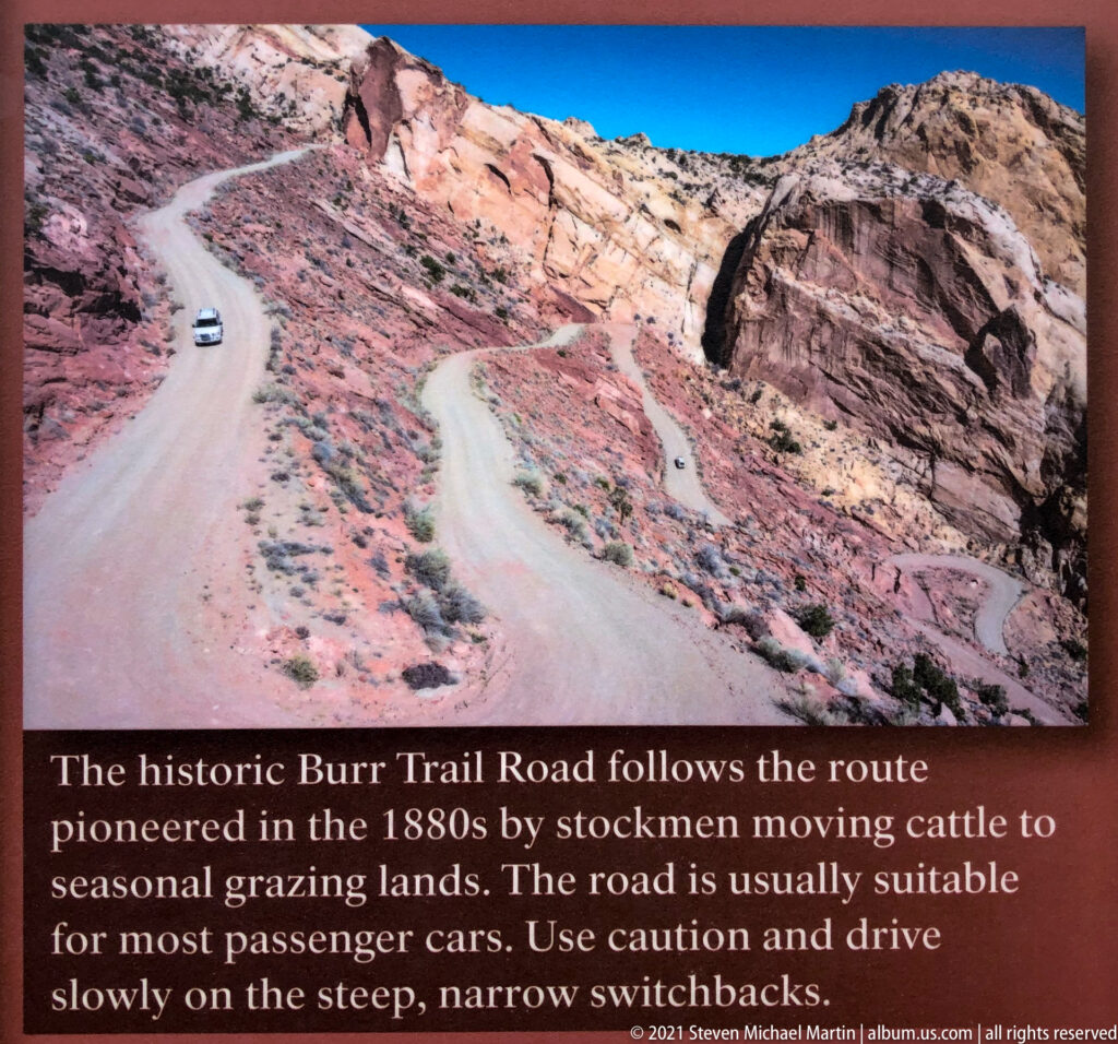 A white vehicle follows a dusty switchback road between red and cream sandstone slopes.
