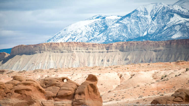 Snow-covered mountain peaks rise beyond pale cliffs and rounded sandstone in the foreground.