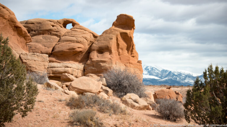 A tall orange sandstone outcrop stands in the foreground, with a snowy mountain visible through the opening to its right.