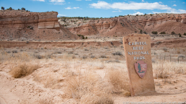 A stone Capitol Reef National Park sign stands in sandy grassland before layered red cliffs.