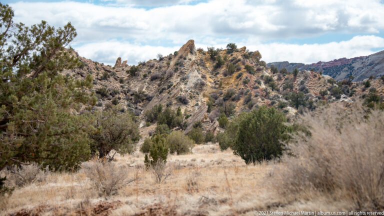 A low rocky ridge with scattered junipers rises above dry grass, with distant cliffs beneath clouds.