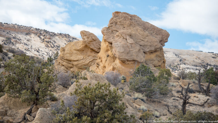 Rounded tan sandstone blocks rise above scattered junipers and shrubs on a pale hillside.