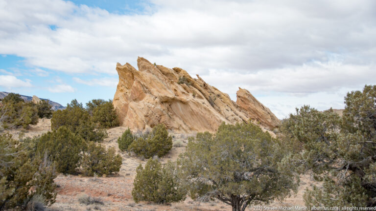 Jagged pale sandstone slabs tilt above dense juniper scrub beneath a cloudy sky.