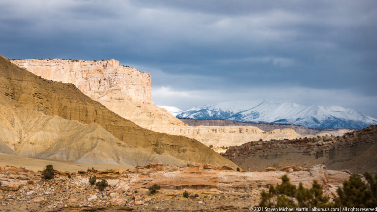 Cream and yellow sedimentary cliffs descend toward a canyon, with snowy mountains under dark clouds beyond.