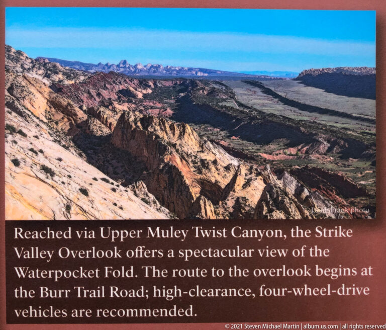 Panoramic valley view with layered red rock ridges and the distant Waterpocket Fold beneath blue sky.