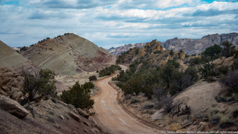 A winding dirt road passes between scrub-covered gray and rust hills toward distant jagged ridges.