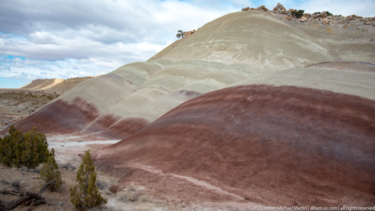 Rounded hills with pale gray, mauve, and rust bands rise above sparse juniper under a cloudy sky.