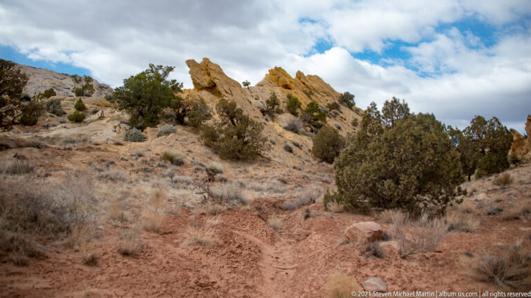 A narrow path climbs toward a jagged sandstone ridge among juniper and dry grasses.