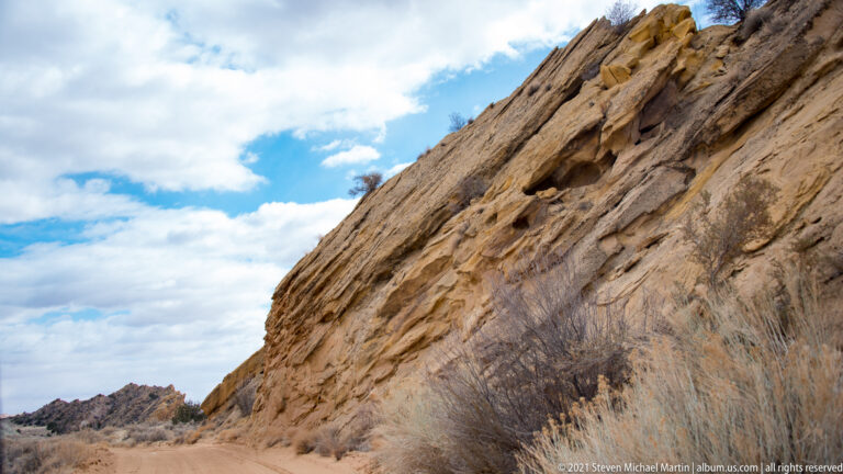 A steep tilted sandstone wall rises beside a dirt track bordered by dry grasses.
