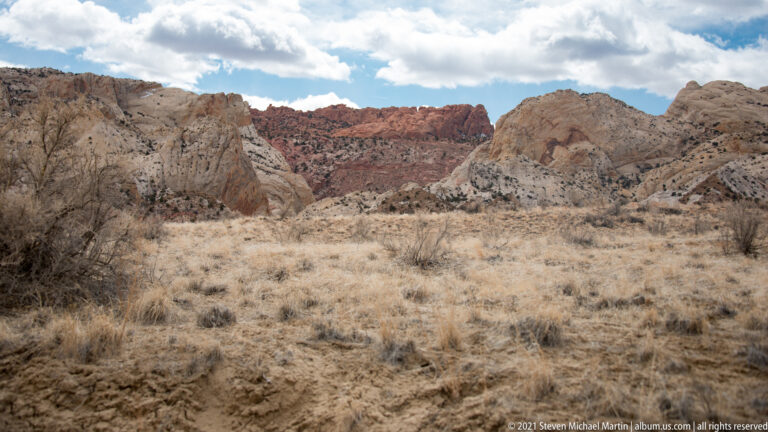 Dry grasses cover an open wash between pale rock walls and a distant red cliff band.