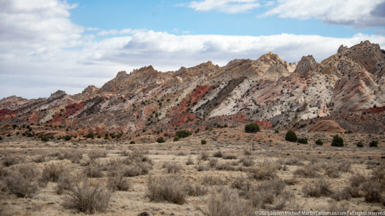 Red, cream, and gray rock bands run across a jagged ridge above a dry shrub plain.