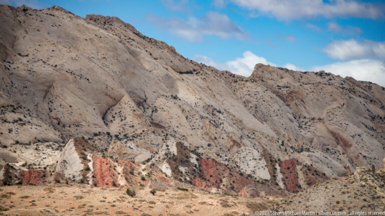 A pale gray eroded slope with shallow gullies descends to red and cream bands.