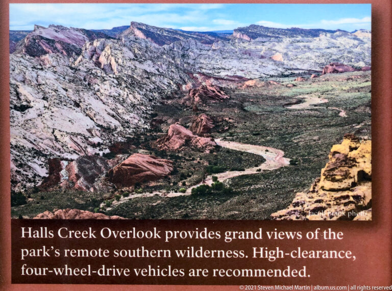 A high overlook frames a winding track through a broad green-brown valley and layered red and cream ridges.