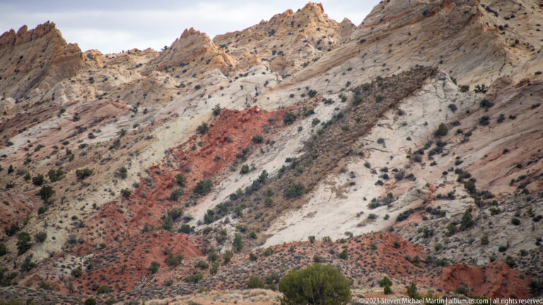 Diagonal red and pale rock bands cross a steep shrub-dotted hillside beneath a cloudy sky.