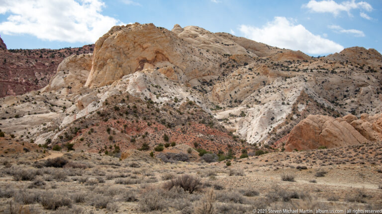 A pale rounded outcrop stands before layered red and cream hills above a dry valley.