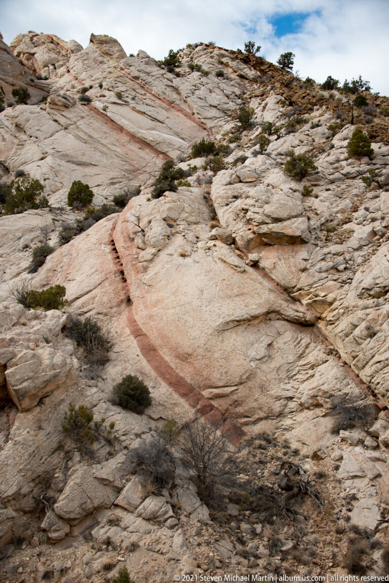 A broad pale sandstone slab slopes downward, crossed by a rust-colored seam and scattered shrubs.